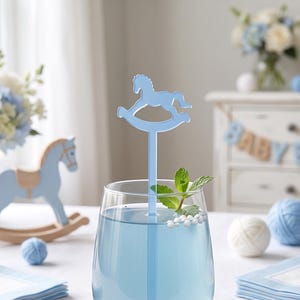 May include: A clear glass tumbler filled with light blue liquid and a light blue stirring stick featuring a rocking horse silhouette at the top. A sprig of fresh green mint and small white spherical garnishes float on the surface of the drink.