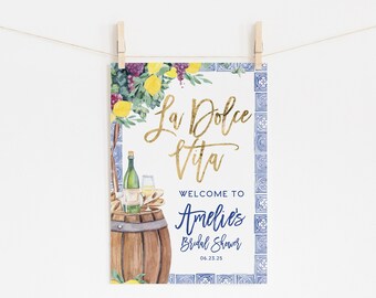 May include: A welcome sign for a bridal shower with a watercolor illustration of a barrel of wine, grapes, lemons, and a blue and white tiled background. The text reads "La Dolce Vita" in gold script and "Welcome to Amelie's Bridal Shower 06.23.25" in blue script.