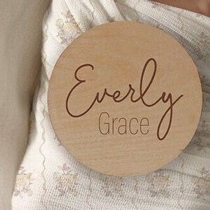 K&ouml;nnte beinhalten: Eine runde Holzplakette mit dem Namen "Everly Grace", der in kursiver Schrift graviert ist.
