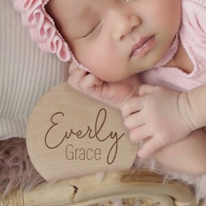 K&ouml;nnte beinhalten: Nahaufnahme eines schlafenden Babys, das eine rosa R&uuml;schenhaube und einen rosa &auml;rmellosen Strampler tr&auml;gt. Ein Holz-Kreis mit dem Text "Everly Grace" befindet sich im Vordergrund.