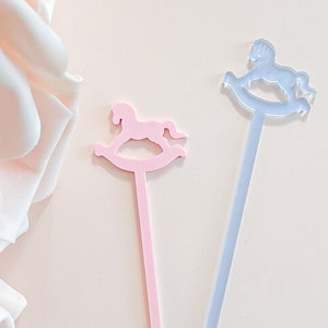 May include: Two long, slender stirrers featuring a rocking horse silhouette at the top. One stirrer is opaque pastel pink, and the other is a translucent light blue. Each has a flat, straight stem ending in a small circular base at the bottom.