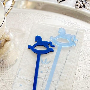 May include: Two long, slender drink stirrers made of acrylic. One stirrer is a deep navy blue and the other is a light sky blue. Each stirrer features a flat, silhouette-style rocking horse design at the top and a small circular base at the bottom.