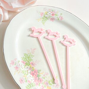 May include: Three light pink stir sticks, each featuring a rocking horse silhouette at the top. The stirrers have a long, slender stem ending in a small circular base. These items are arranged diagonally on a white plate with a floral pattern.