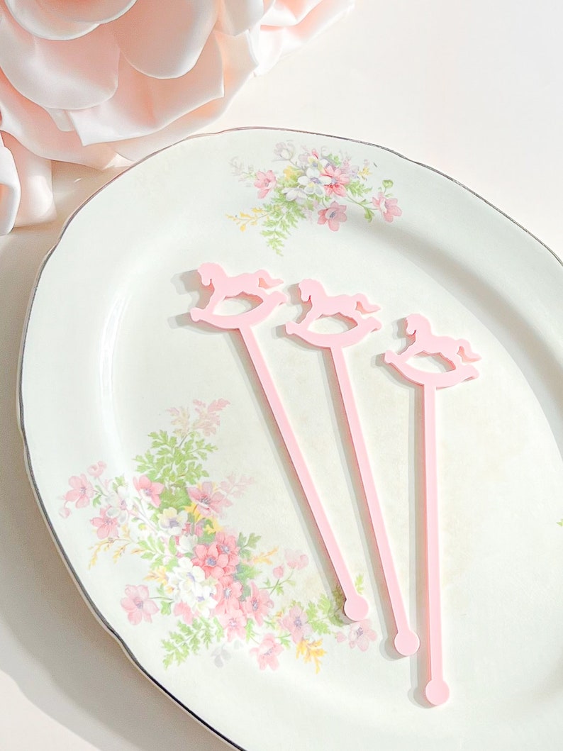 May include: Three light pink stir sticks, each featuring a rocking horse silhouette at the top. The stirrers have a long, slender stem ending in a small circular base. These items are arranged diagonally on a white plate with a floral pattern.