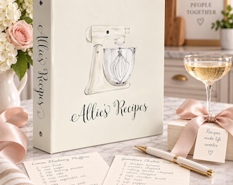 May include: A white three-ring binder featuring a cream-colored stand mixer illustration and the text "Allie's Recipes" on the cover and spine. Two recipe cards with handwritten text and a white and gold pen are placed in front of the binder.