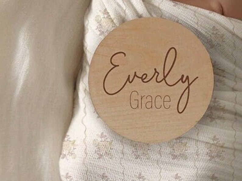 K&ouml;nnte beinhalten: Eine runde Holzplakette mit dem Namen "Everly Grace", der in kursiver Schrift graviert ist.