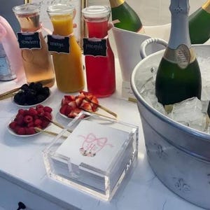 May include: A mimosa bar setup featuring three glass carafes filled with strawberry, orange, and pineapple juices, each with a black chalkboard label. A silver bucket holds champagne bottles in ice, alongside small plates of fresh berries and napkins.