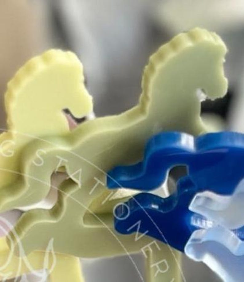 May include: A set of flat, horse-shaped acrylic pieces in pale yellow and translucent blue. The pieces feature a smooth, die-cut silhouette of a horse in a galloping pose. A faint, circular watermark with text is overlaid across the objects.