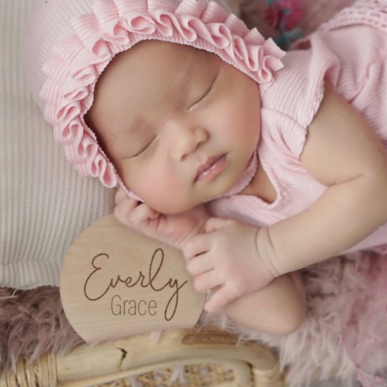 K&ouml;nnte beinhalten: Nahaufnahme eines schlafenden Babys, das eine rosa R&uuml;schenhaube und einen rosa &auml;rmellosen Strampler tr&auml;gt. Ein Holz-Kreis mit dem Text "Everly Grace" befindet sich im Vordergrund.