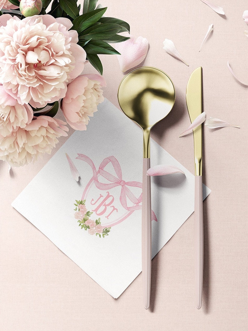 May include: A gold and light pink spoon and knife set placed next to a white square napkin featuring a pink ribbon, floral wreath, and the letters JBr. A bouquet of light pink peonies with green leaves is positioned in the upper left corner.