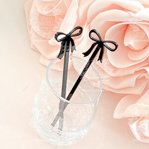 May include: Two black acrylic drink stirrers with bow accents rest in a clear glass. The stirrers are positioned against a soft pink floral background, creating an elegant and stylish presentation. The glass is partially filled with liquid.