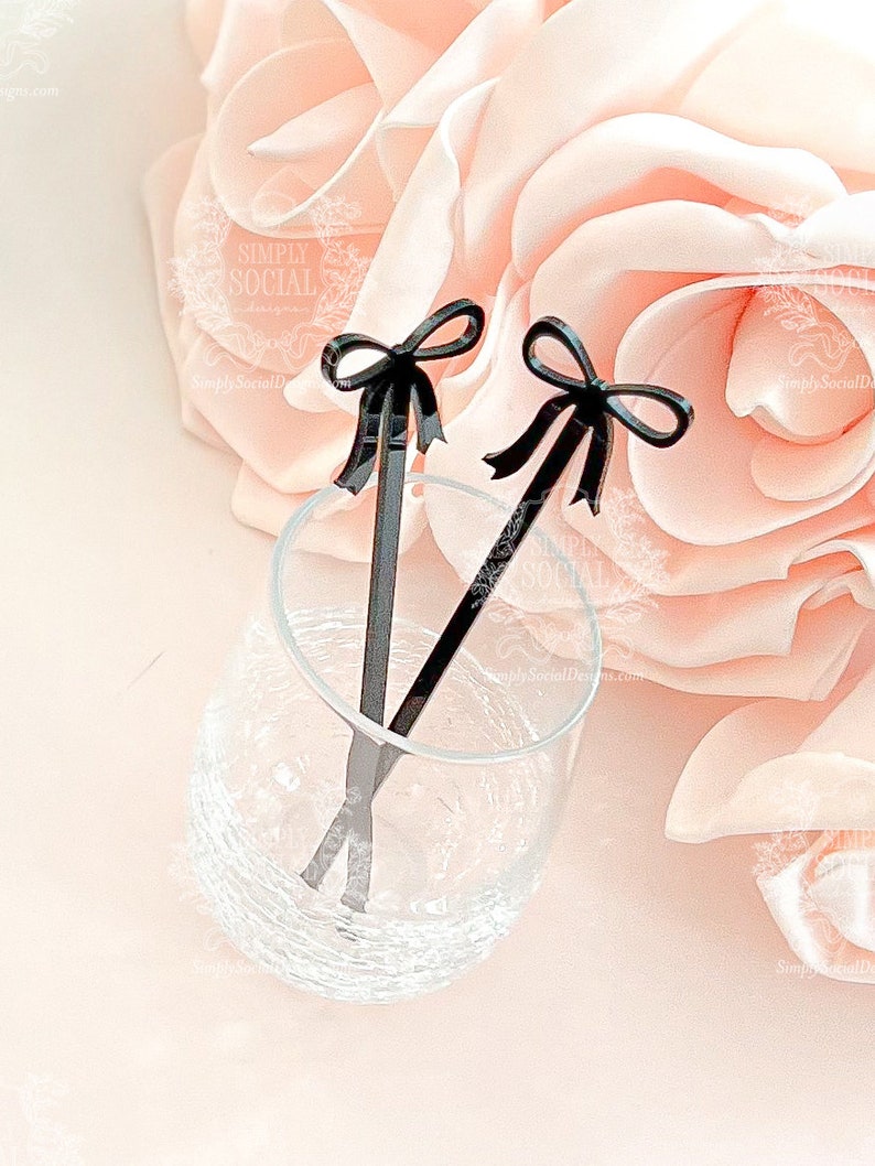 May include: Two black acrylic drink stirrers with bow accents rest in a clear glass. The stirrers are positioned against a soft pink floral background, creating an elegant and stylish presentation. The glass is partially filled with liquid.