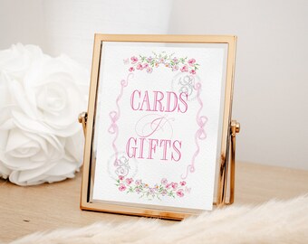 May include: A gold-framed sign with a white background and pink floral accents. The sign reads "Cards & Gifts" in pink lettering.