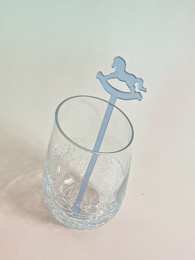 May include: A clear glass tumbler with a crackled texture containing a light blue acrylic drink stirrer. The stirrer features a flat, silhouette-style rocking horse topper at the top end, extending vertically from the base of the glass.