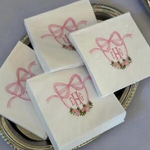 May include: A stack of square white paper napkins featuring a printed design of a pink ribbon tied in a bow above a floral wreath. The center of the wreath contains the monogram letters EHG in a pink script font, printed on each napkin.