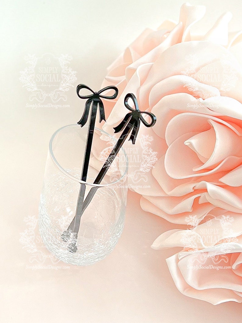 May include: Two black stir sticks with bow-shaped tops rest in a clear glass. The glass sits on a light pink surface next to a large, soft, pink flower. The stir sticks are a decorative accessory.
