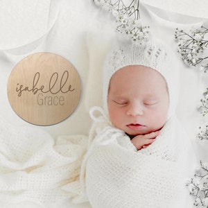 K&ouml;nnte beinhalten: Ein rundes Holzschild mit dem Namen "Isabelle Grace", der in braunen Buchstaben eingraviert ist. Ein Neugeborenes schl&auml;ft in einer wei&szlig;en gestrickten Decke und M&uuml;tze.