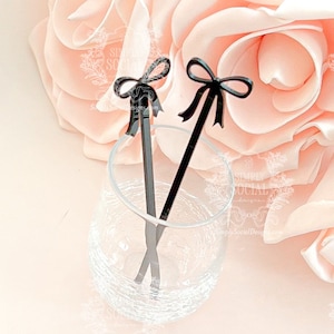 May include: Two black cocktail stirrers with bow-shaped tops are displayed in a clear glass. The stirrers are set against a soft pink background with a large rose. The stirrers add a touch of elegance to any drink.