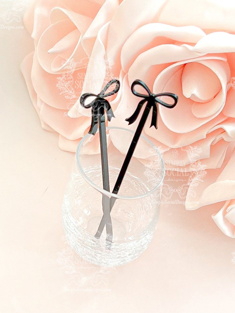 May include: Two black cocktail stirrers with bow-shaped tops are displayed in a clear glass. The stirrers are set against a soft pink background with a large rose. The stirrers add a touch of elegance to any drink.
