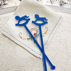 May include: Two blue plastic drink stirrers, each featuring a flat silhouette of a rocking horse at the top and a circular end at the base. The stirrers are positioned in a crisscross pattern over a square, off-white linen napkin with embroidery.