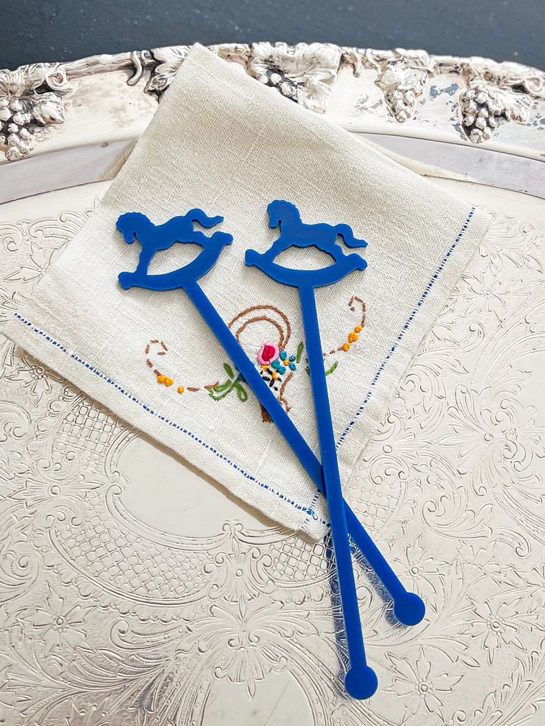 May include: Two blue plastic drink stirrers, each featuring a flat silhouette of a rocking horse at the top and a circular end at the base. The stirrers are positioned in a crisscross pattern over a square, off-white linen napkin with embroidery.