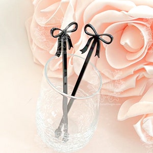 May include: Two black acrylic drink stirrers with bow-shaped tops rest in a clear glass. The stirrers are set against a backdrop of soft pink roses. The stirrers are a decorative addition to cocktails and other beverages.
