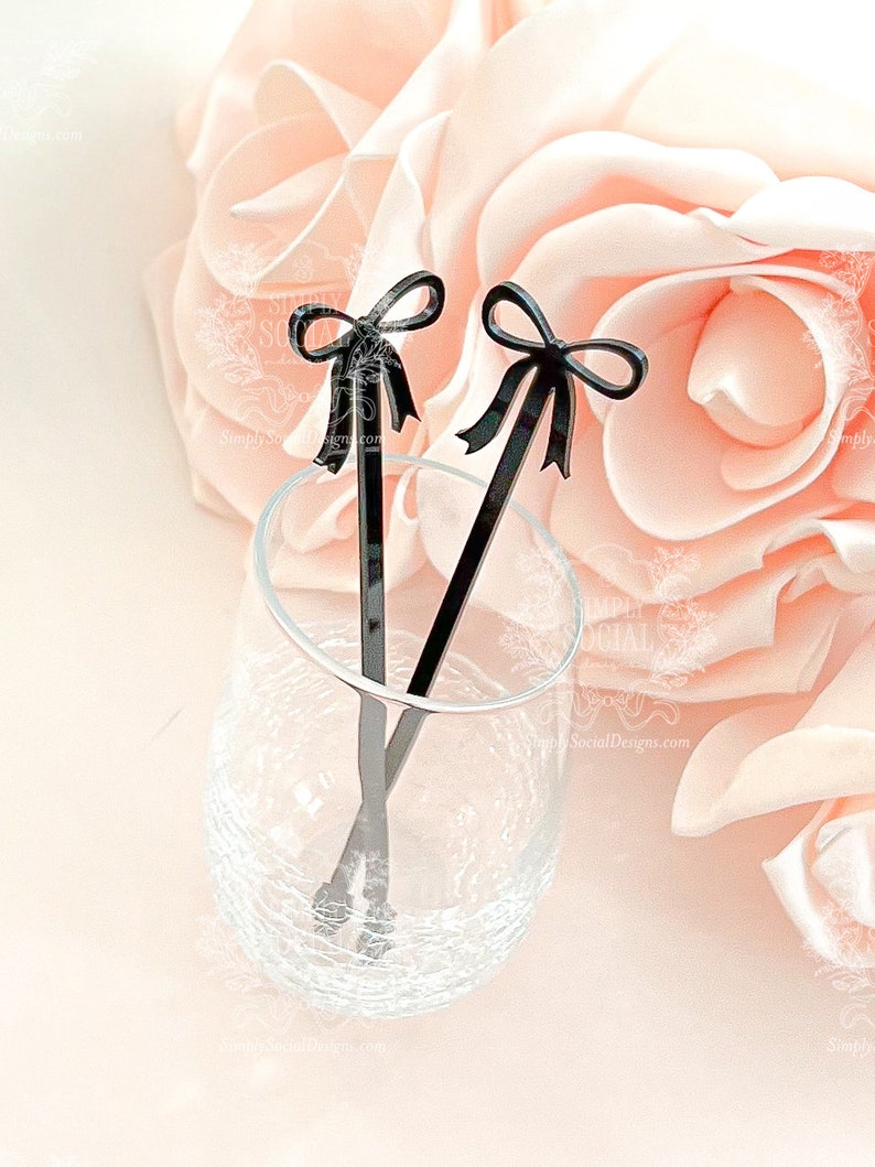 May include: Two black acrylic drink stirrers with bow-shaped tops rest in a clear glass. The stirrers are set against a backdrop of soft pink roses. The stirrers are a decorative addition to cocktails and other beverages.