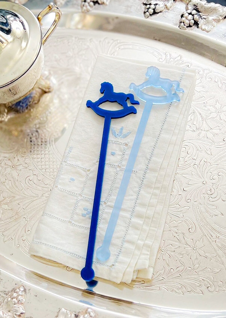 May include: Two long, slender drink stirrers made of acrylic. One stirrer is a deep navy blue and the other is a light sky blue. Each stirrer features a flat, silhouette-style rocking horse design at the top and a small circular base at the bottom.