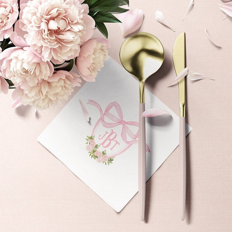 May include: A gold and matte light pink spoon and knife set placed next to a white square napkin featuring a pink ribbon bow, a floral wreath, and the monogram JBT, accompanied by a cluster of light pink peony flowers and scattered petals.
