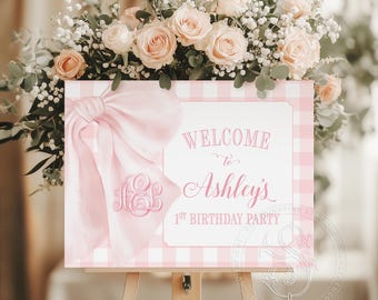 May include: A pink and white sign on an easel, with the text "WELCOME to Ashley's 1st BIRTHDAY PARTY". The sign features a pink bow and a monogram. The sign is decorated with a pink gingham pattern and is surrounded by peach roses and greenery.