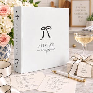 May include: A white three-ring binder with a black bow graphic and the text Olivia's Recipes on the front and spine. Two recipe cards with handwritten text, a white pen with gold accents, and a stack of cards tied with a ribbon are present.