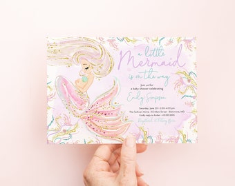 May include: A baby shower invitation with a mermaid illustration. The invitation is for Emily Simpson and features a pink and purple watercolor background with gold glitter accents. The text reads "a little mermaid is on the way".