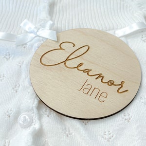 K&ouml;nnte beinhalten: Eine runde Holzplakette mit dem Namen "Eleanor Jane", der in einer kursivschriftlichen Schriftart graviert ist.