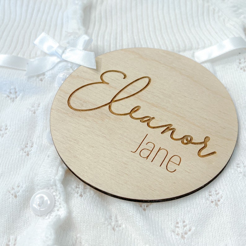 K&ouml;nnte beinhalten: Eine runde Holzplakette mit dem Namen "Eleanor Jane", der in einer kursivschriftlichen Schriftart graviert ist.