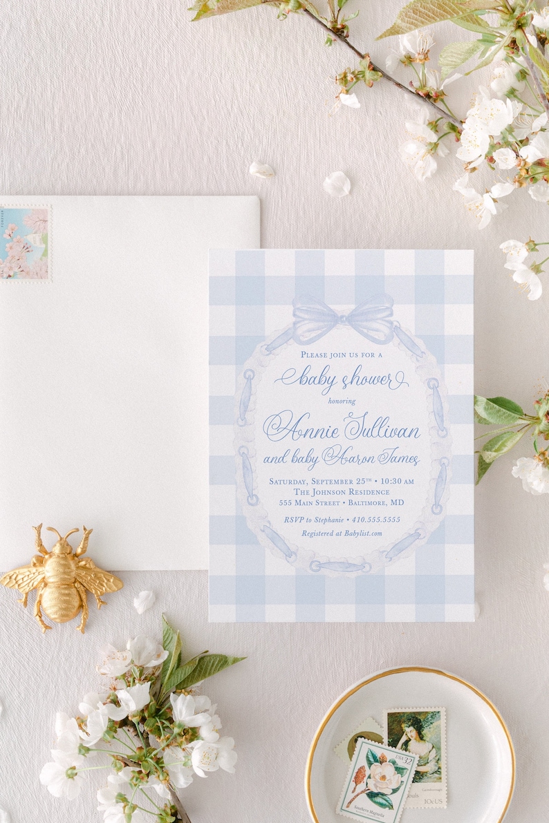 May include: A blue and white gingham baby shower invitation with a lace design and a bow. The invitation reads "Please join us for a baby shower honoring Annie Sullivan and baby Aaron James. Saturday, September 25th, 10:30 AM. The Johnson Residence, 555 Main Street, Baltimore, MD. RSVP to Stephanie 410.555.5555. Registered at Babylist.com"