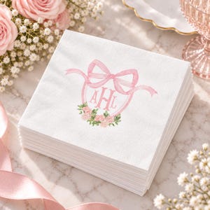 May include: A stack of square white paper napkins featuring a printed design of a pink watercolor bow tied around a circular frame. Inside the frame are the letters AHL in a script font, with a small cluster of pink flowers and green leaves below.