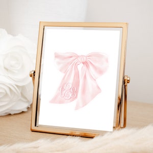 May include: A pink watercolor bow with a white monogram in the center. The bow is in a gold frame.