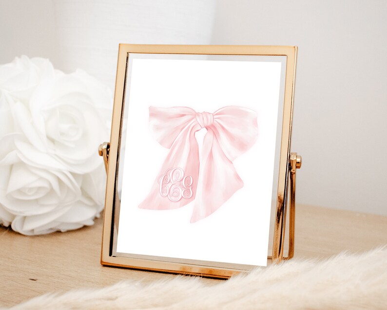 May include: A pink watercolor bow with a white monogram in the center. The bow is in a gold frame.