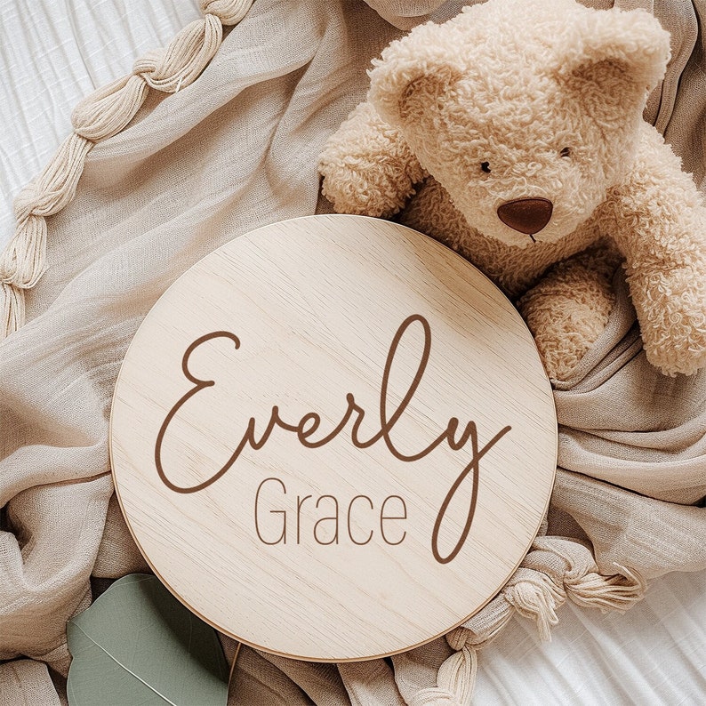 K&ouml;nnte beinhalten: Ein rundes Holzschild mit dem Namen "Everly Grace" in brauner Kursivschrift. Das Schild liegt auf einer beigefarbenen Decke mit einem Teddyb&auml;ren.