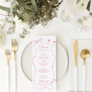 May include: A baby brunch table setting with a menu, cutlery, and floral decorations. The menu, in pink script, reads "Elena's Baby Brunch." Gold forks, knife, and spoon with white handles are arranged on a white plate and napkin.