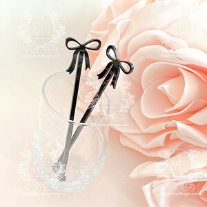 May include: Two black stir sticks with bow-shaped tops rest in a clear glass. The glass sits on a light pink surface next to a large, soft, pink flower. The stir sticks are a decorative accessory.