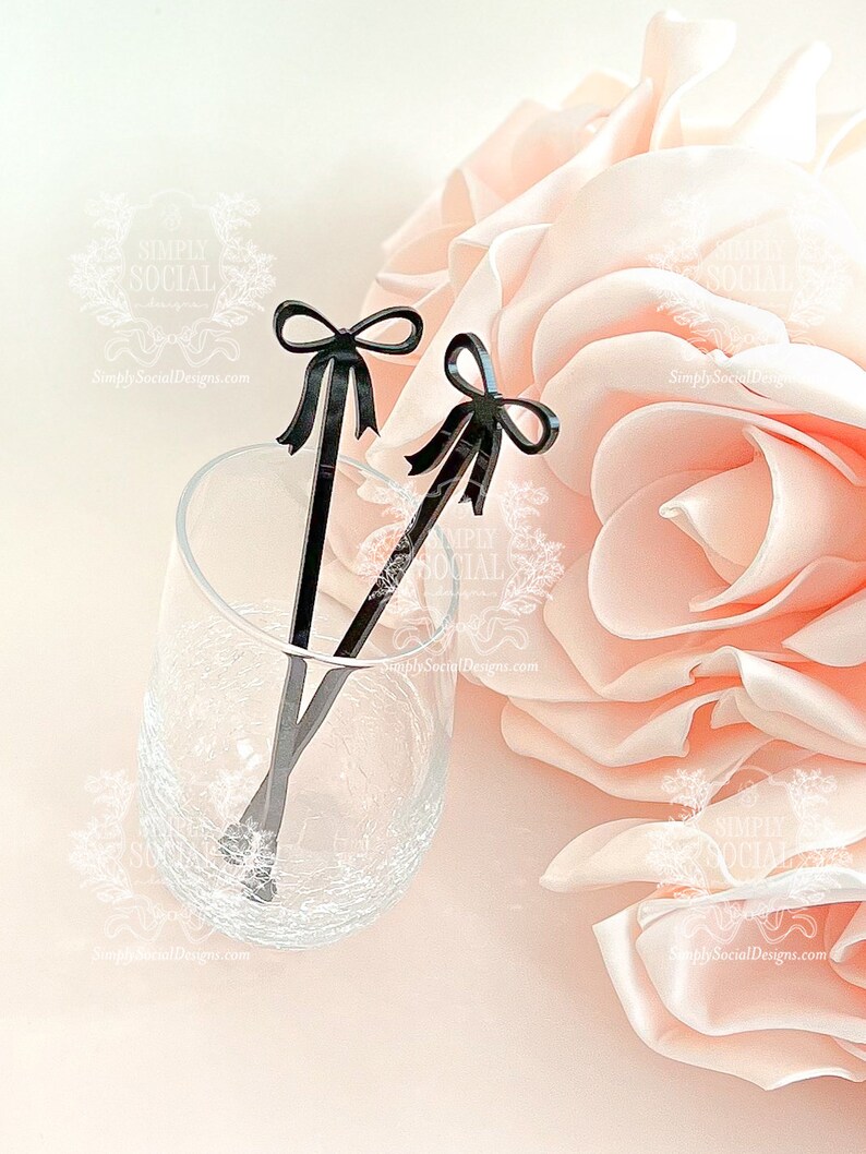 May include: Two black stir sticks with bow-shaped tops rest in a clear glass. The glass sits on a light pink surface next to a large, soft, pink flower. The stir sticks are a decorative accessory.