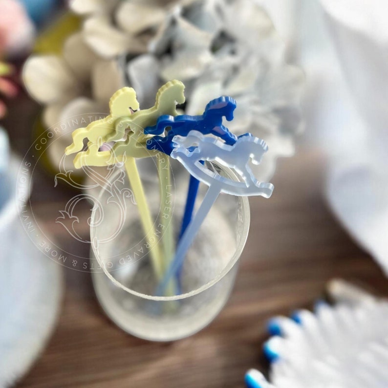 May include: Three acrylic drink stirrers in a clear glass, each featuring a rocking horse topper. The stirrers are in pale yellow, dark blue, and translucent light blue. A circular watermark with text is visible over the stirrers.
