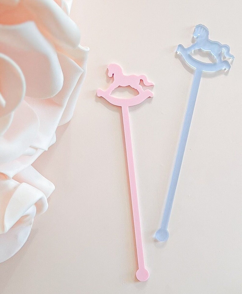 May include: Two long, slender stirrers featuring a rocking horse silhouette at the top. One stirrer is opaque pastel pink, and the other is a translucent light blue. Each has a flat, straight stem ending in a small circular base at the bottom.