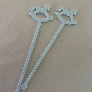 May include: Two light gray stirrers, each featuring a flat, cutout design of a rocking horse at the top and a small circular base at the bottom. The stirrers are long, slender, and uniform in color, with a smooth, matte finish throughout the structure.
