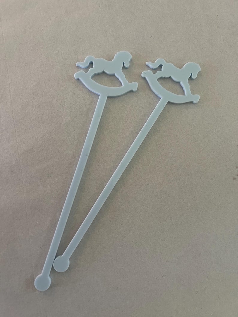 May include: Two light gray stirrers, each featuring a flat, cutout design of a rocking horse at the top and a small circular base at the bottom. The stirrers are long, slender, and uniform in color, with a smooth, matte finish throughout the structure.