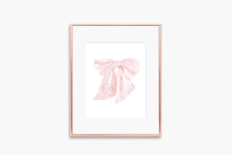 May include: A pink satin bow with a monogram in the center. The bow is tied in a simple knot and the ends are left long. The bow is framed in a gold frame.