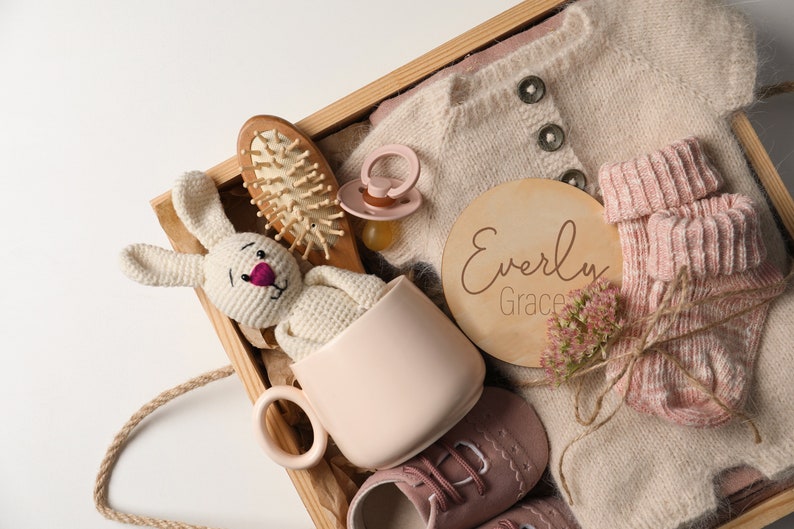 K&ouml;nnte beinhalten: Eine Holzkiste gef&uuml;llt mit Babyartikeln, darunter ein gestrickter Pullover, Socken, ein Schnuller, eine Tasse, ein geh&auml;keltes H&auml;schen, eine Haarb&uuml;rste und ein Holzschild mit dem Namen "Everly Grace".