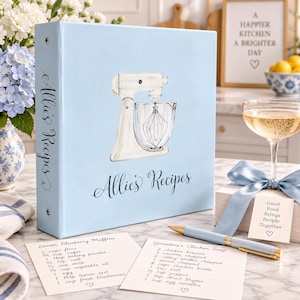May include: A light blue three-ring binder with the text Allie's Recipes and a white stand mixer illustration on the cover. Two recipe cards for Lemon Blueberry Muffins and Grandma's Chicken Soup, a light blue pen, and a gift tag are included.
