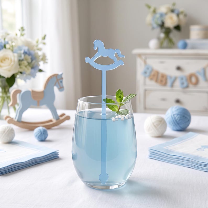 May include: A clear glass tumbler filled with light blue liquid and a light blue stirring stick featuring a rocking horse silhouette at the top. A sprig of fresh green mint and small white spherical garnishes float on the surface of the drink.